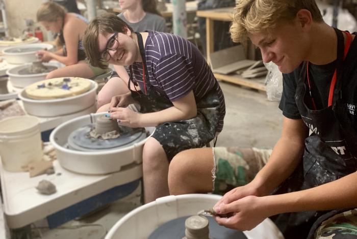Four teens are sitting at a row of pottery wheels. They all have clay in front of them. One of them is looking and smiling at the camera. 